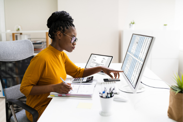 lady sitting at desk pointing at screen with rows of information. Writing down on paper.