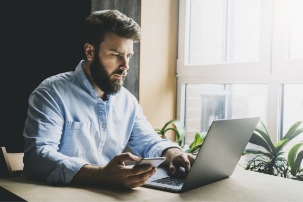 Man sitting focused on a Notebook Computer sitting on table with Cell Phone in hand.