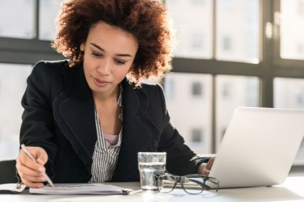 Lady sitting while doing work on both Notebook computer and writing down information on paper.