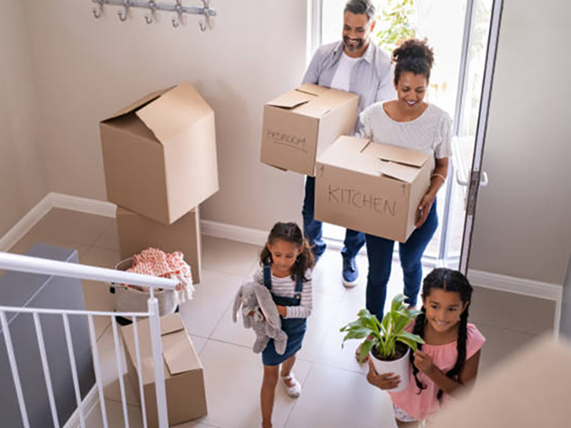 Ethnic family with two children carrying boxes and plant in new home on moving day. High angle view of happy smiling daughters helping mother and father with cardboard boxes in new house. Top view of excited kids having fun walking up stairs running to their rooms while parents holding boxes.